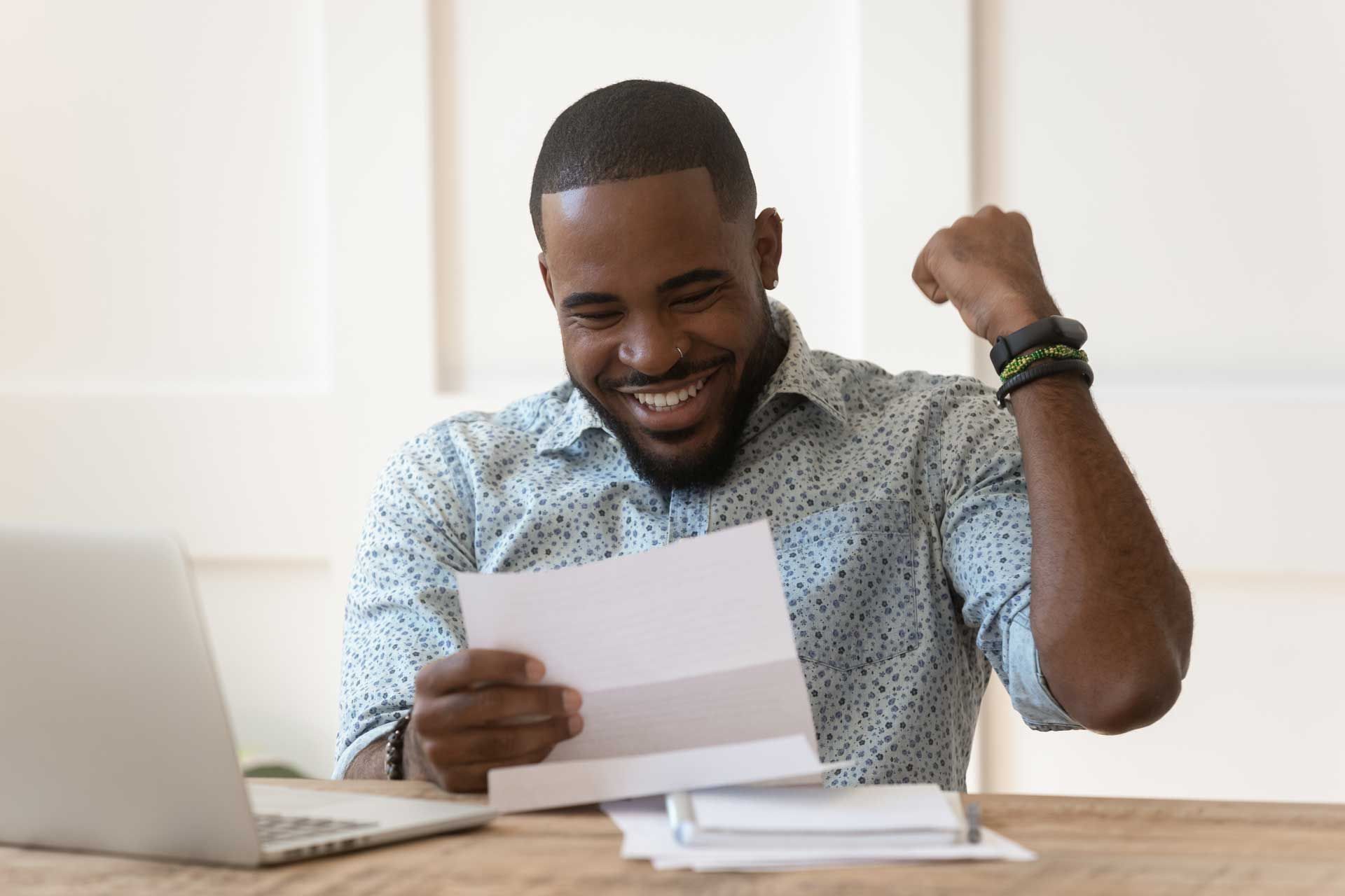 Man reading paper, smiling, fist raised in excitement; laptop on desk.