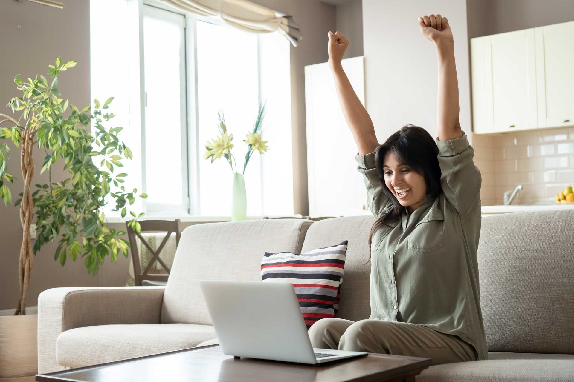 Woman raises arms in celebration, sitting on a sofa with a laptop in a living room.
