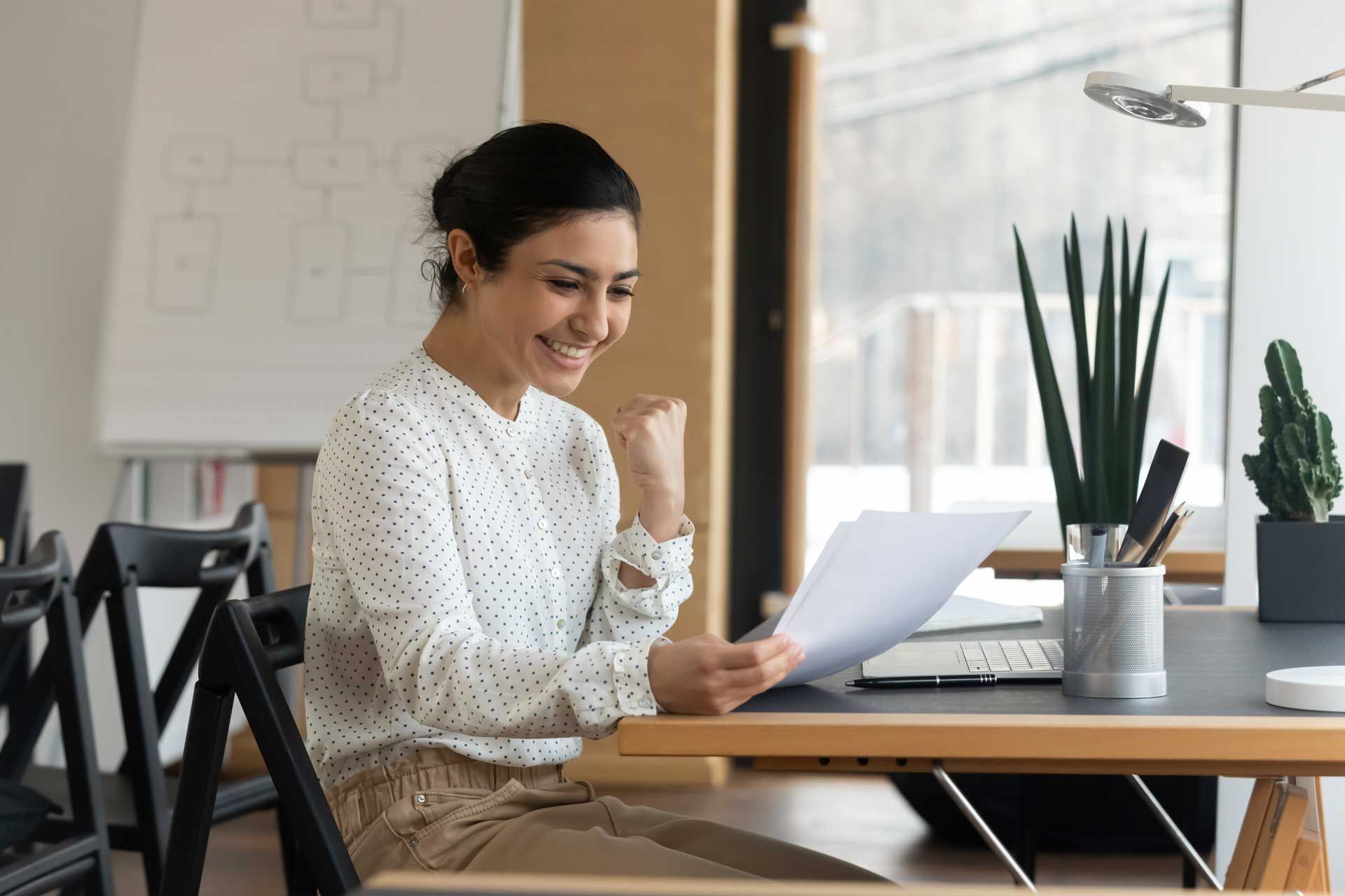 Woman smiles, raises fist, reading paper at desk, celebrating success in office setting.