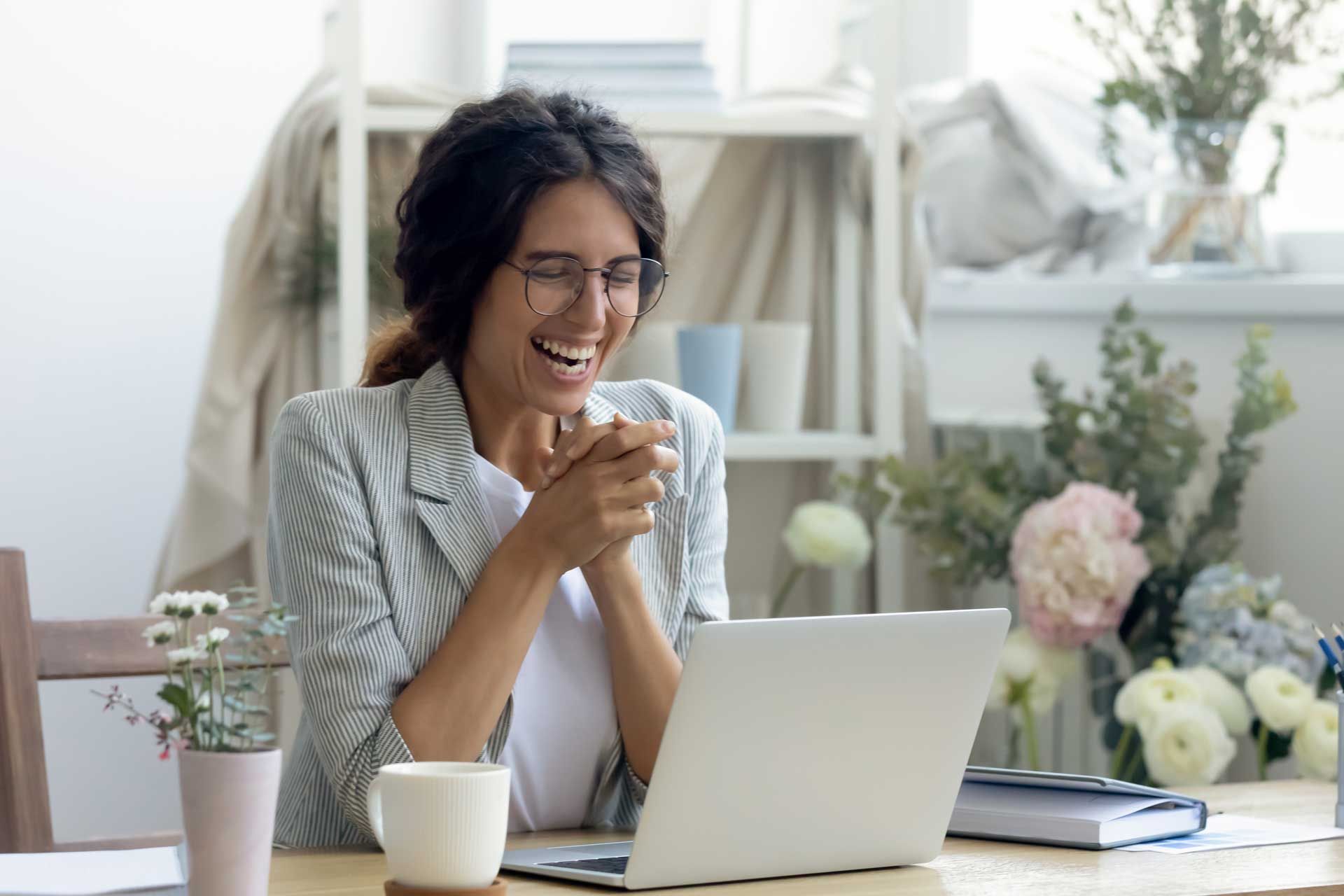 Woman wearing glasses, laughing while looking at laptop. Holding a mug, sitting at a desk with flowers in a bright room.