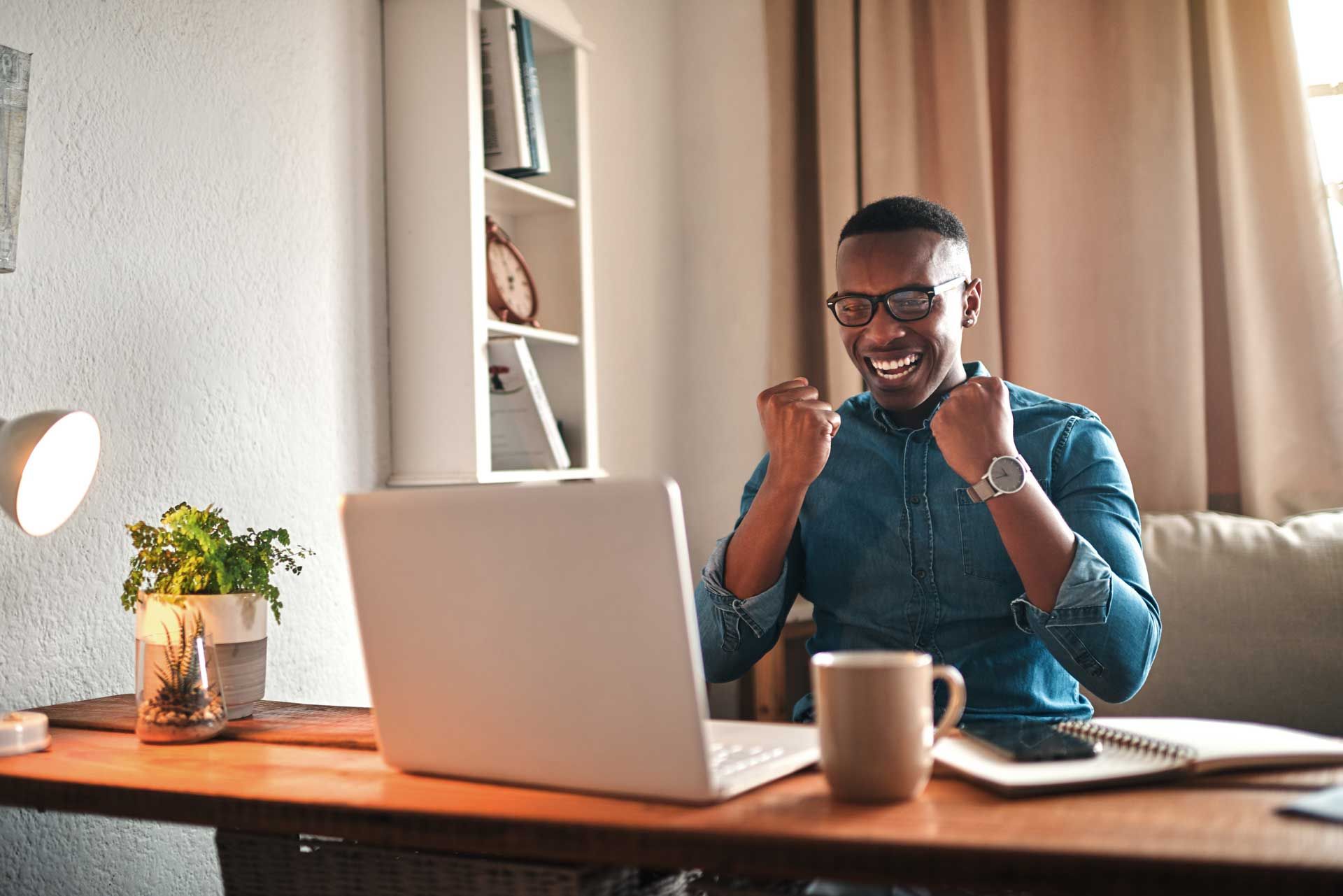 Man with glasses smiles, pumps fists while looking at laptop on desk. White bookshelf in background.