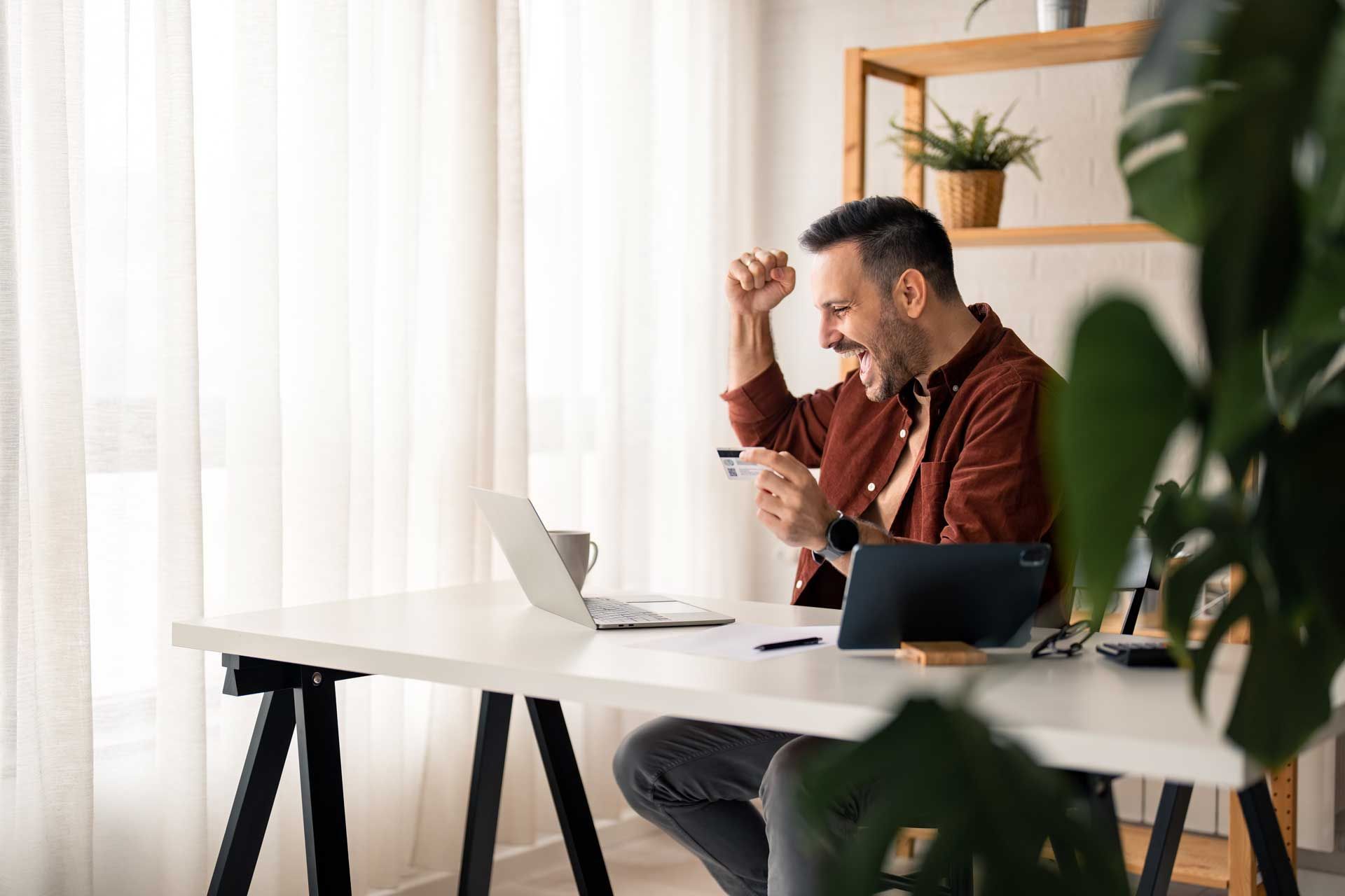 Man celebrating at a desk with laptop, holding a credit card, near a window and plants.