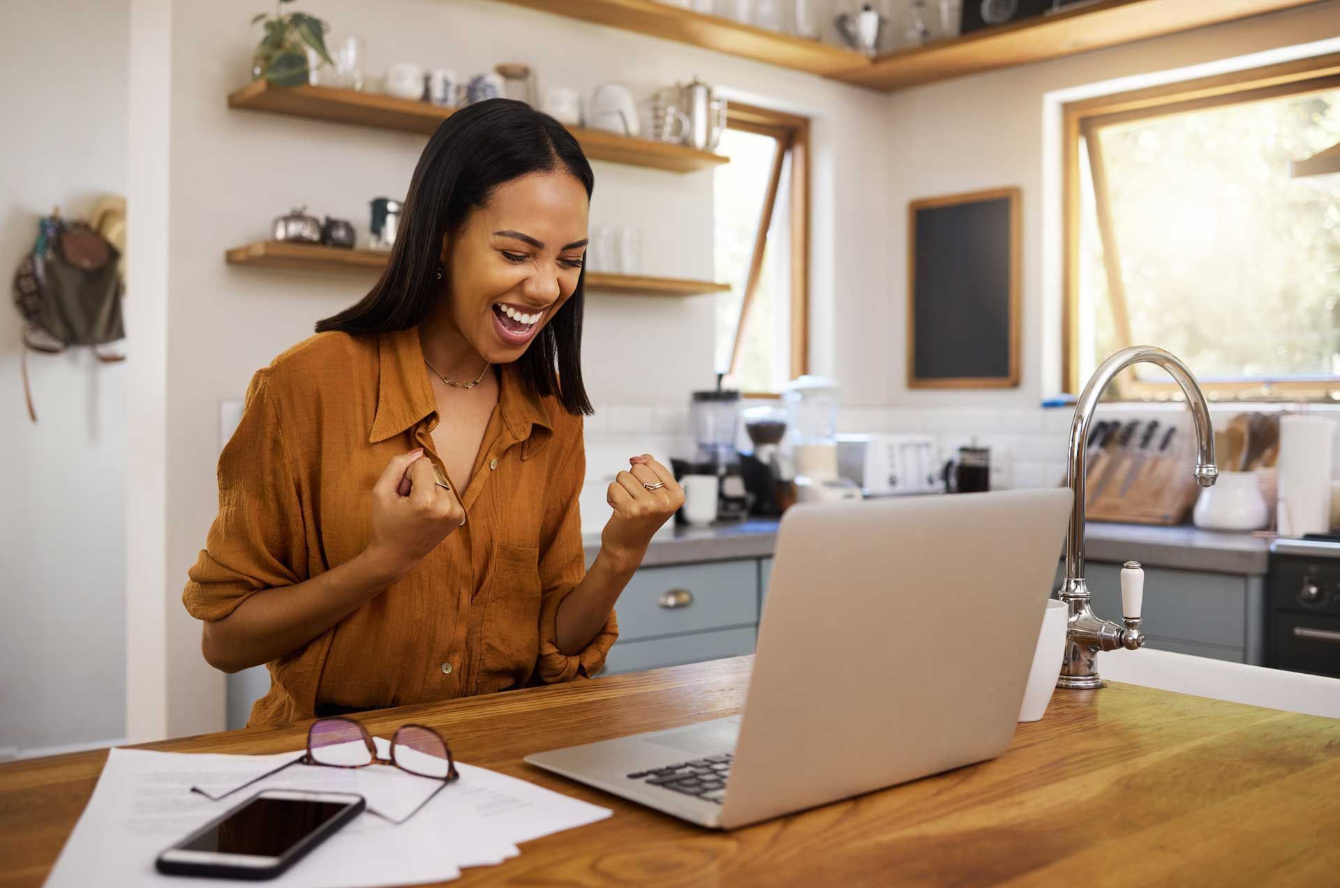 Woman celebrates in kitchen with laptop, fists raised.