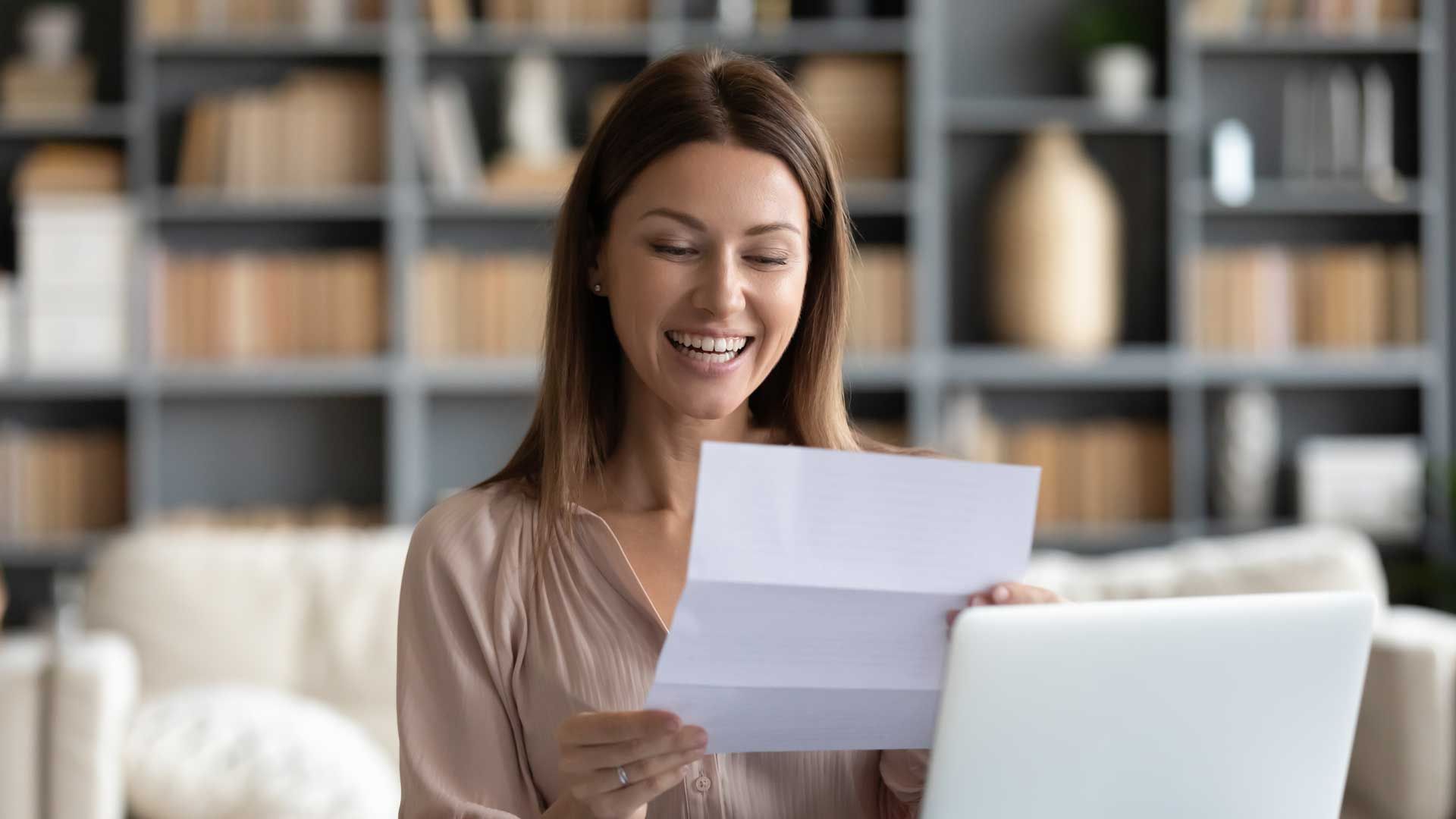 Woman smiling, reading a letter indoors with a laptop on a table.