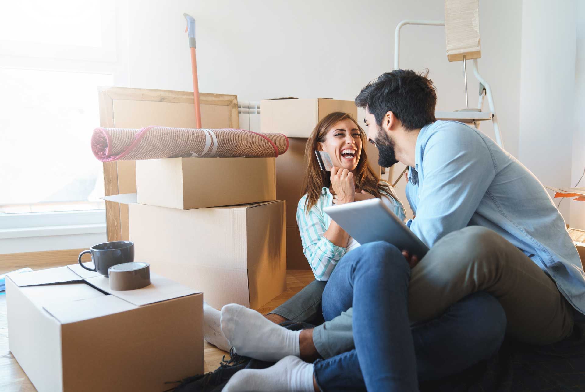 Couple laughs while using a tablet in a room filled with moving boxes.
