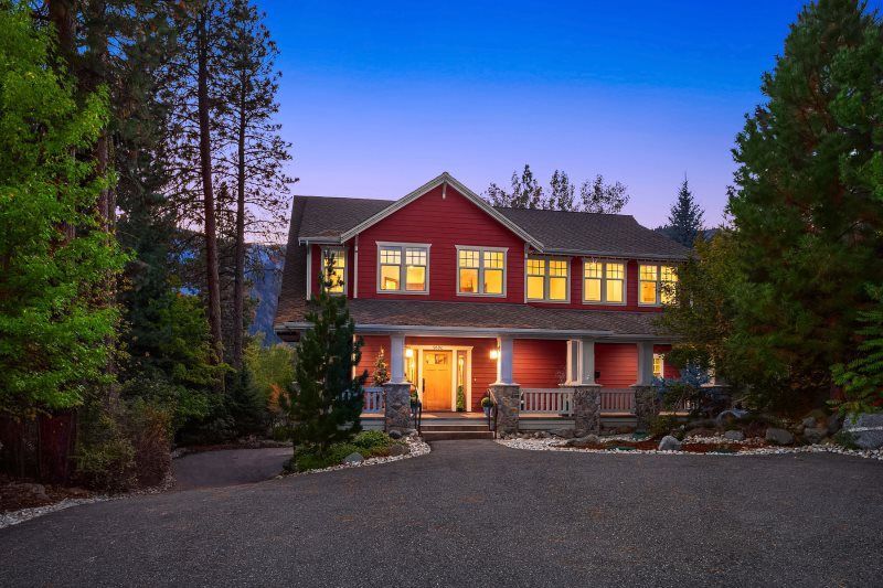 Large two-story house with lit windows and porch at dusk, surrounded by trees and a gravel driveway.