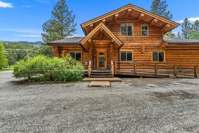 Rustic log cabin with wooden porch and gravel driveway, surrounded by trees and hills under a clear sky