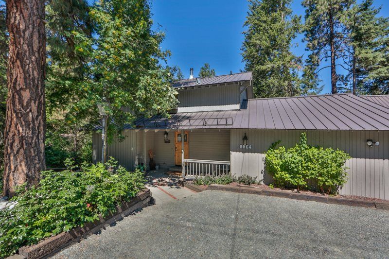 Wooded home exterior with driveway, white railing, and metal roof under clear blue sky