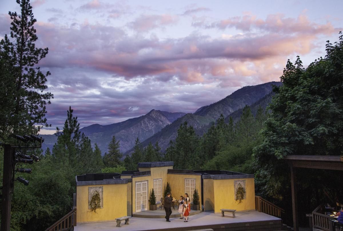 Small yellow cabin with people on a patio at dusk, surrounded by pine trees and mountain peaks.