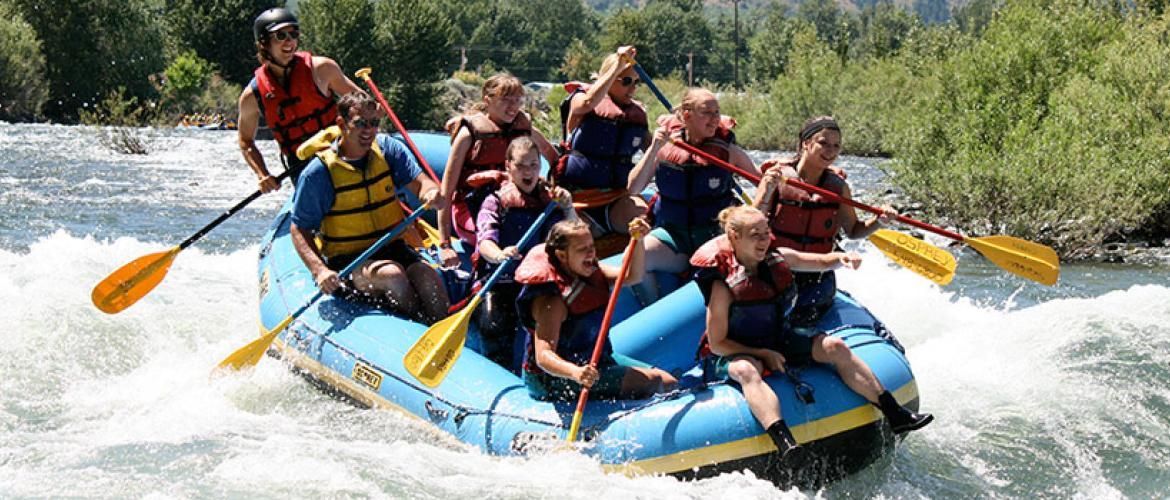 Group whitewater rafting in blue raft, paddling through splashing rapids with yellow oars.