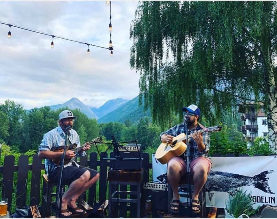Two musicians playing guitar outdoors on a deck, with mountains and greenery in the background.