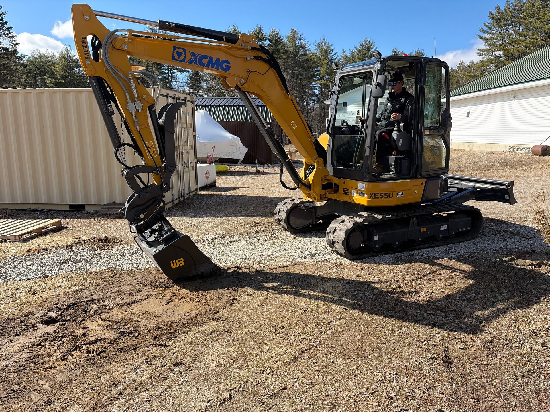 A yellow XCMG excavator with a grapple attachment parked on gravel outdoors near a shipping container.