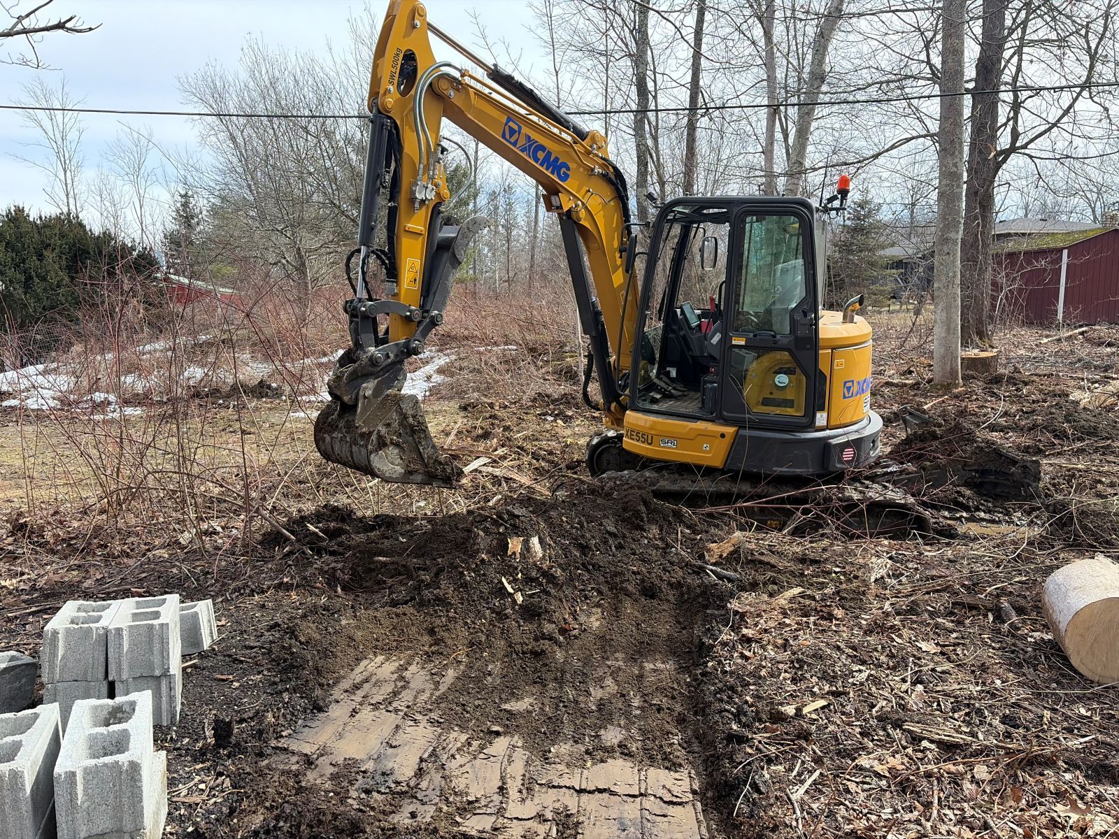 A yellow excavator sits in a wooded area, digging into the earth near several stacked concrete blocks.