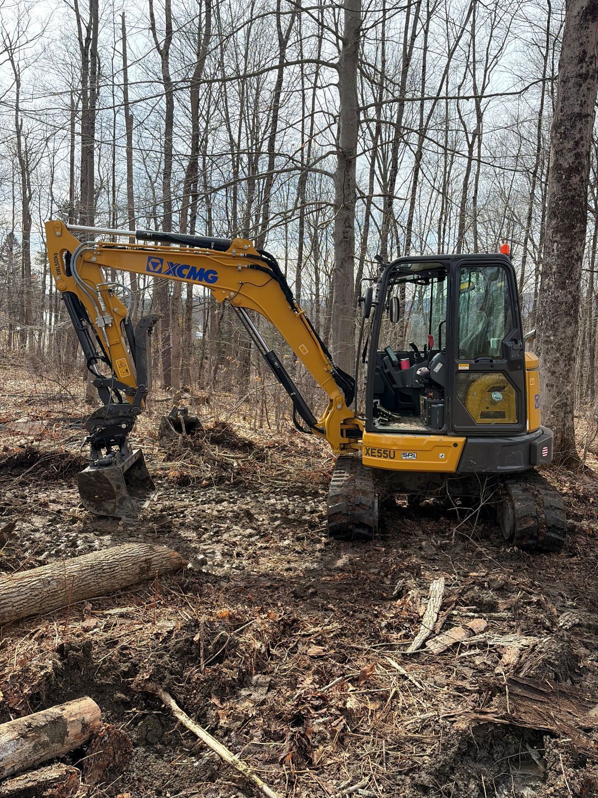 A yellow XCMG excavator parked in a wooded area with bare trees and cleared ground.