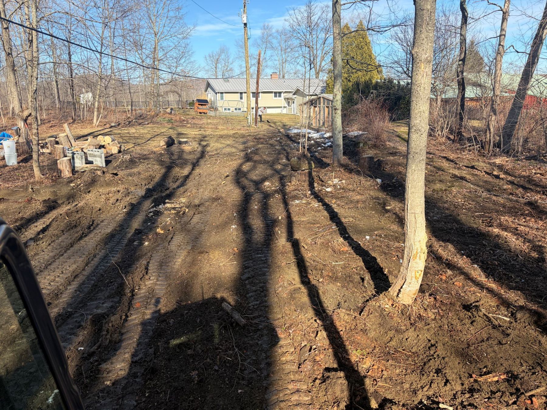 A view from a vehicle looking out onto a freshly cleared, muddy dirt patch in a wooded area with a house in the distance.