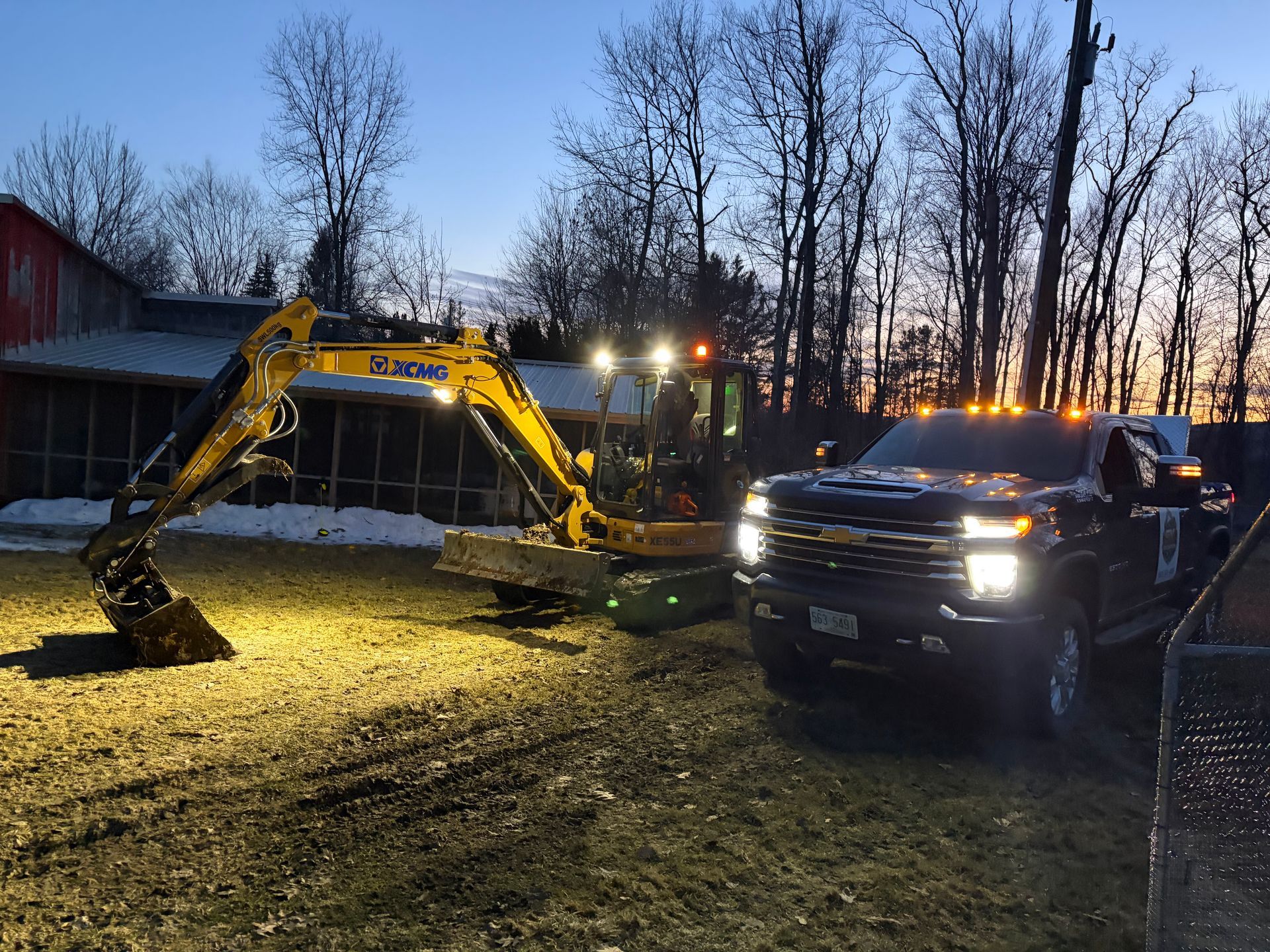 A dark pickup truck with bright headlights parked next to a yellow excavator in a grassy, partially snow-covered field.