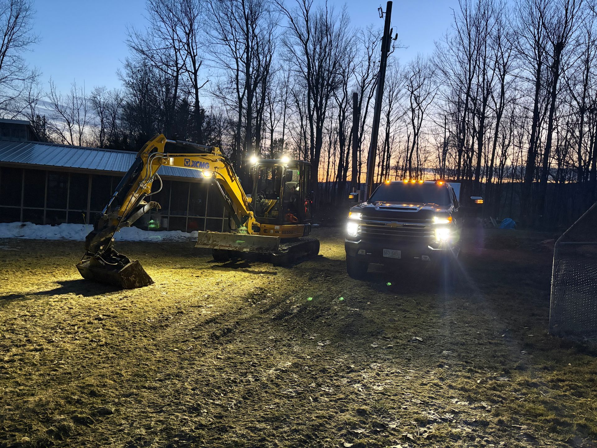 A yellow excavator and a pickup truck with bright headlights illuminate a dirt yard at dusk, backed by bare trees.