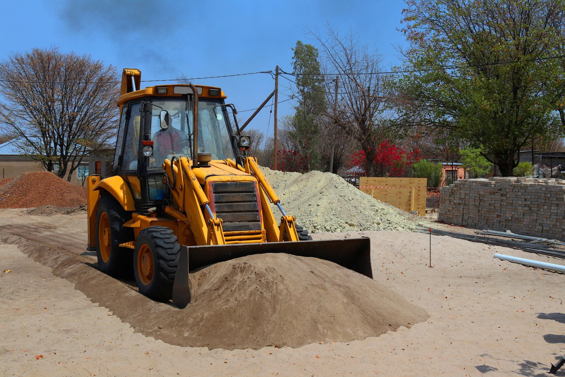 A yellow backhoe loader sits on a sandy construction site with piles of dirt and trees in the background under a blue sky.