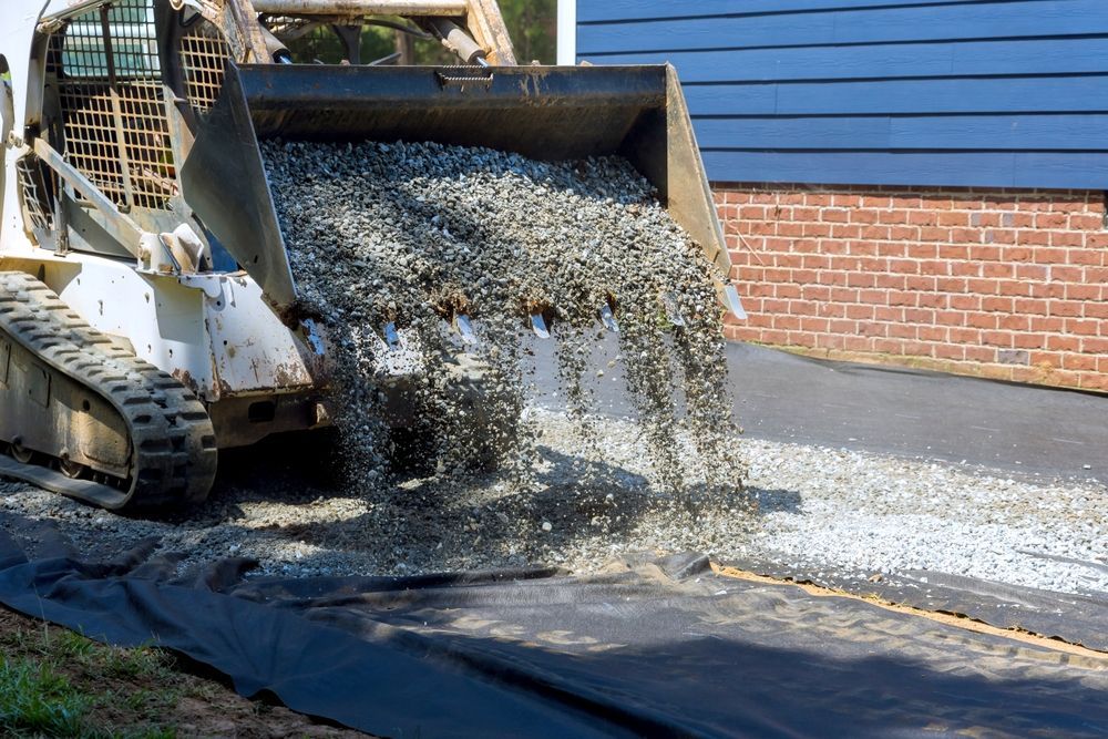 A skid steer loader dumps gray gravel onto a black landscape fabric sheet next to a brick building.