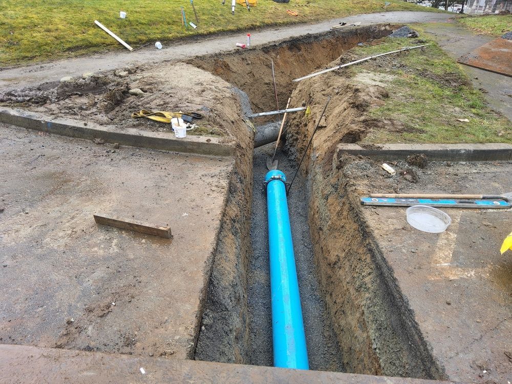 A blue PVC pipe installed in a deep, excavated dirt trench cutting through a concrete surface.