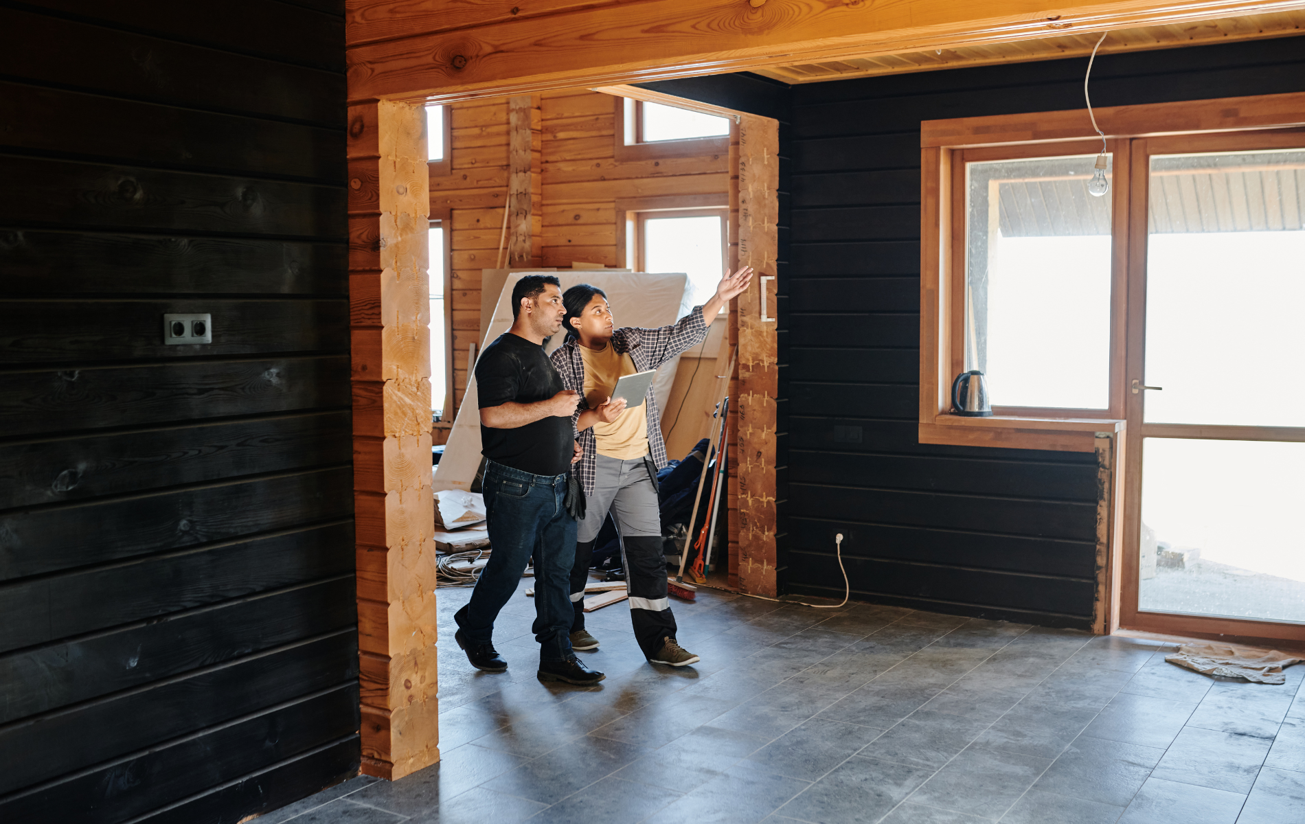 Two people measuring a room under renovation with exposed wood framing and sunlight through windows