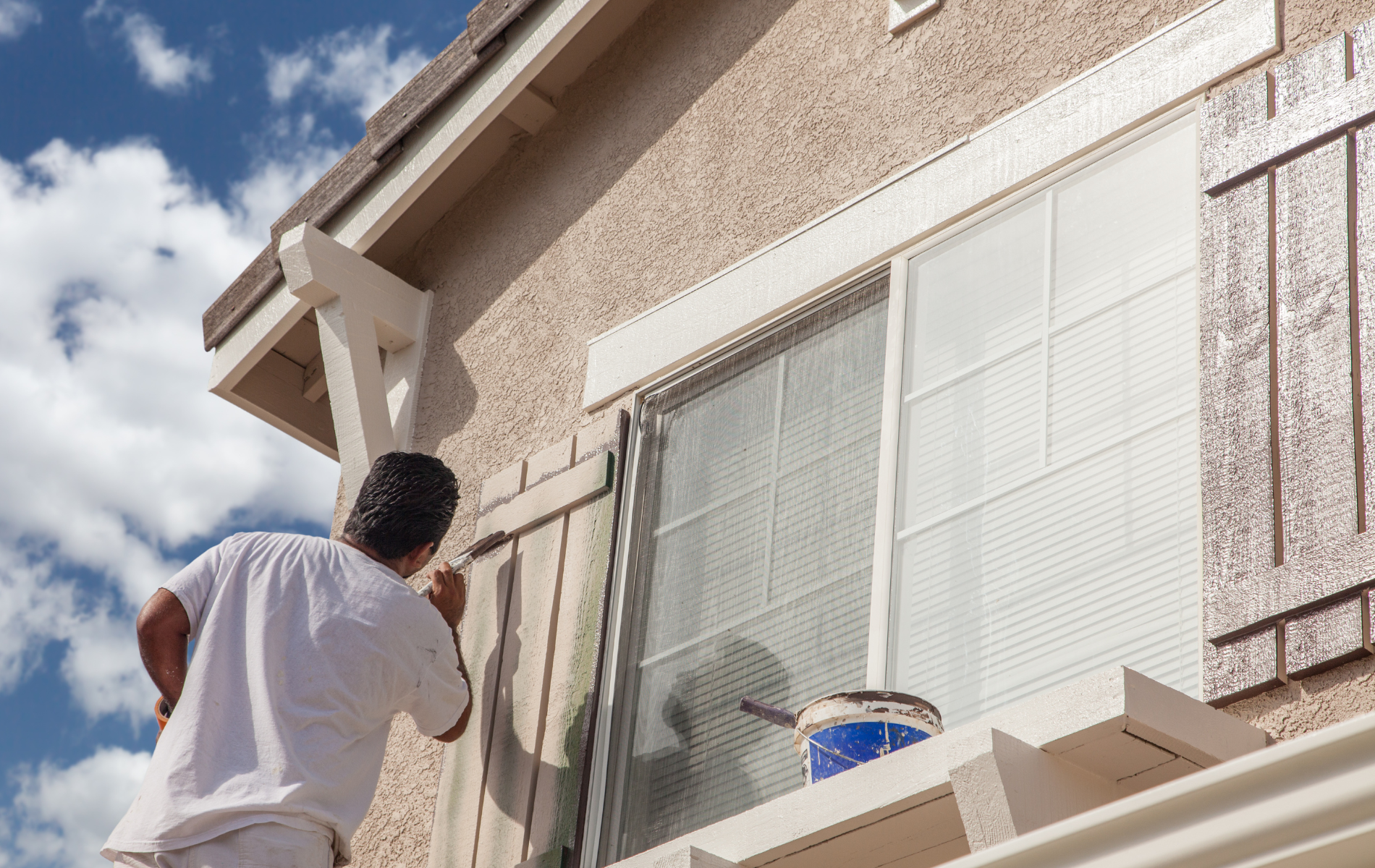 Person painting wooden shutter on a house, blue sky background.
