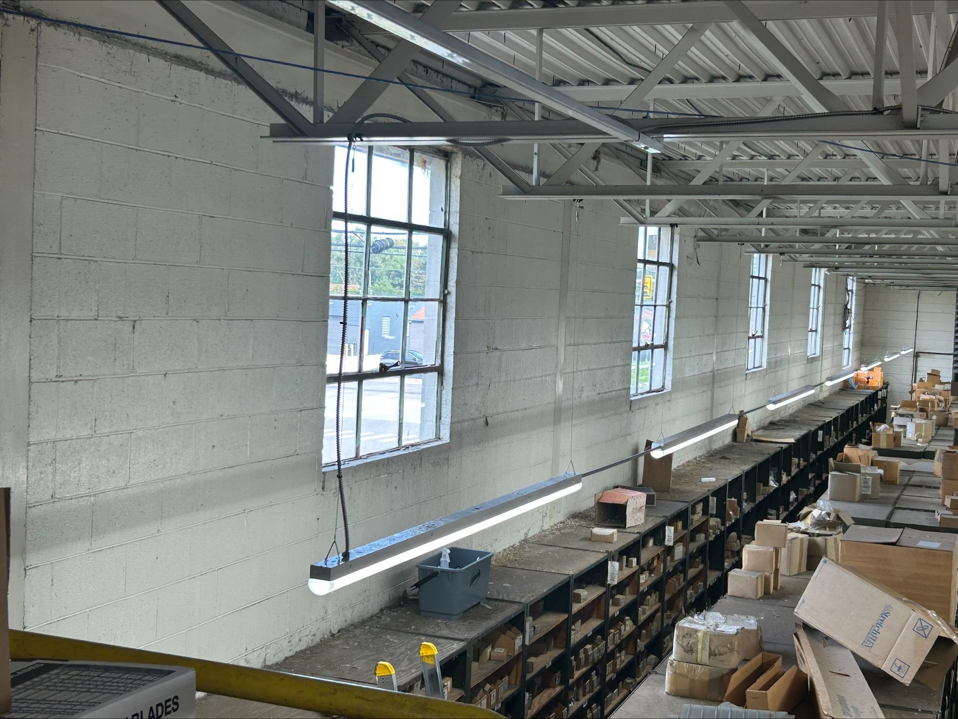 A long industrial storage room with white brick walls, multiple windows, fluorescent lighting, and rows of cluttered shelves.