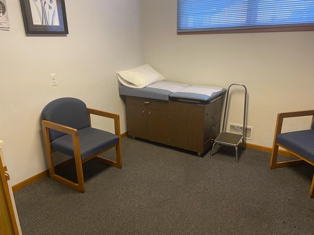 A medical examination room with two blue chairs, an exam table with a pillow, and a small metal step stool.