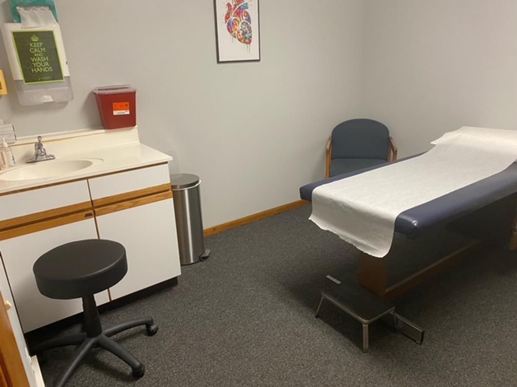 A clinical examination room with a patient bed, a stool, a sink, and a framed anatomical heart poster on the wall.
