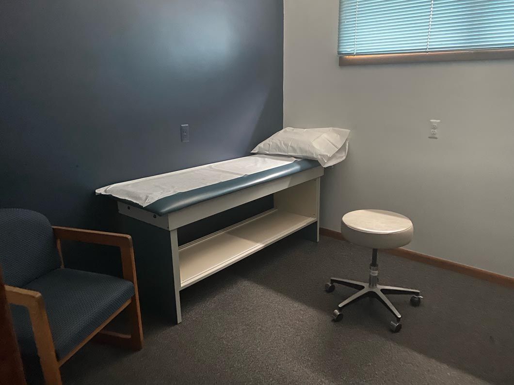 A small clinical exam room with a dark blue accent wall, a padded examination table with paper, a chair, and a rolling stool.