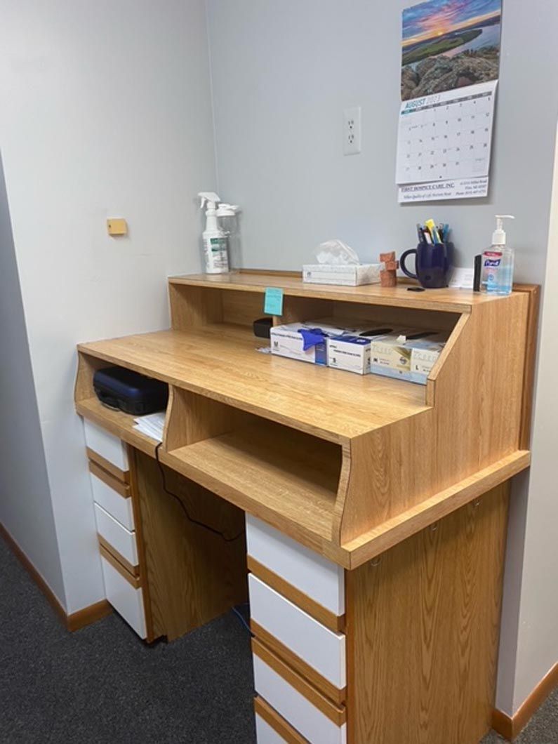 A light wood office desk with white drawers, holding a tissue box, hand sanitizer, spray bottle, and medical supplies.