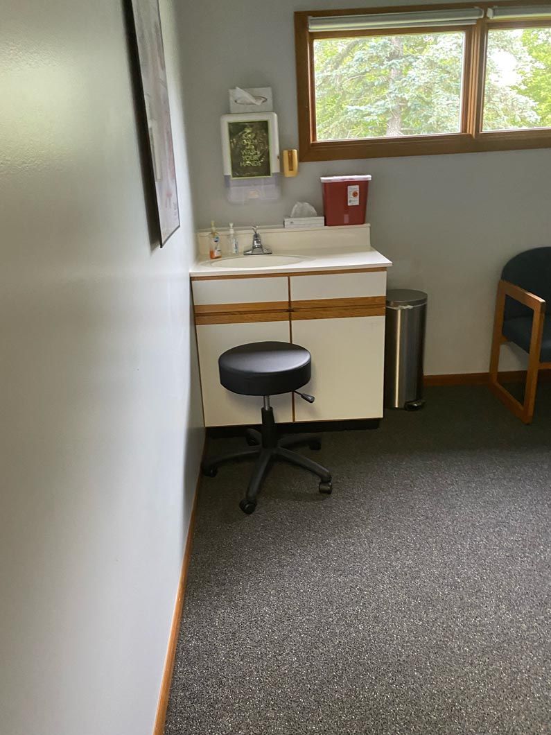 A medical examination room with a rolling stool, white cabinet with a sink, a red sharps container, and a metal trash bin.