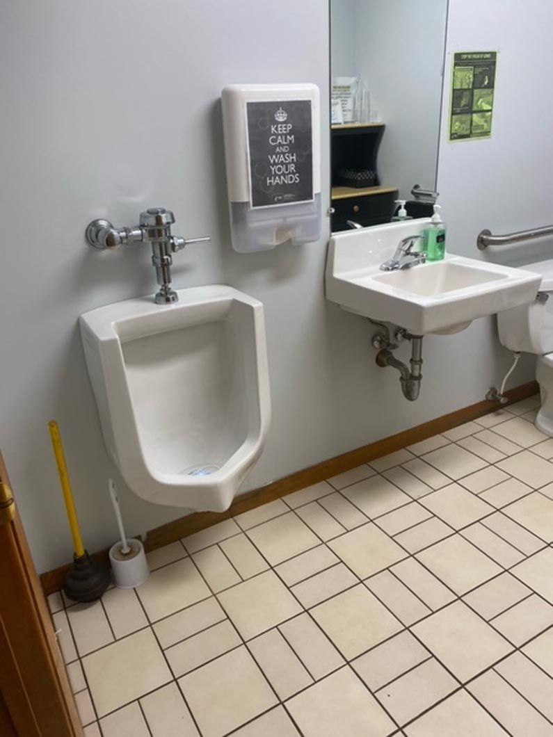 A public restroom featuring a wall-mounted urinal, a sink with a soap dispenser, and a toilet plunger on the floor.