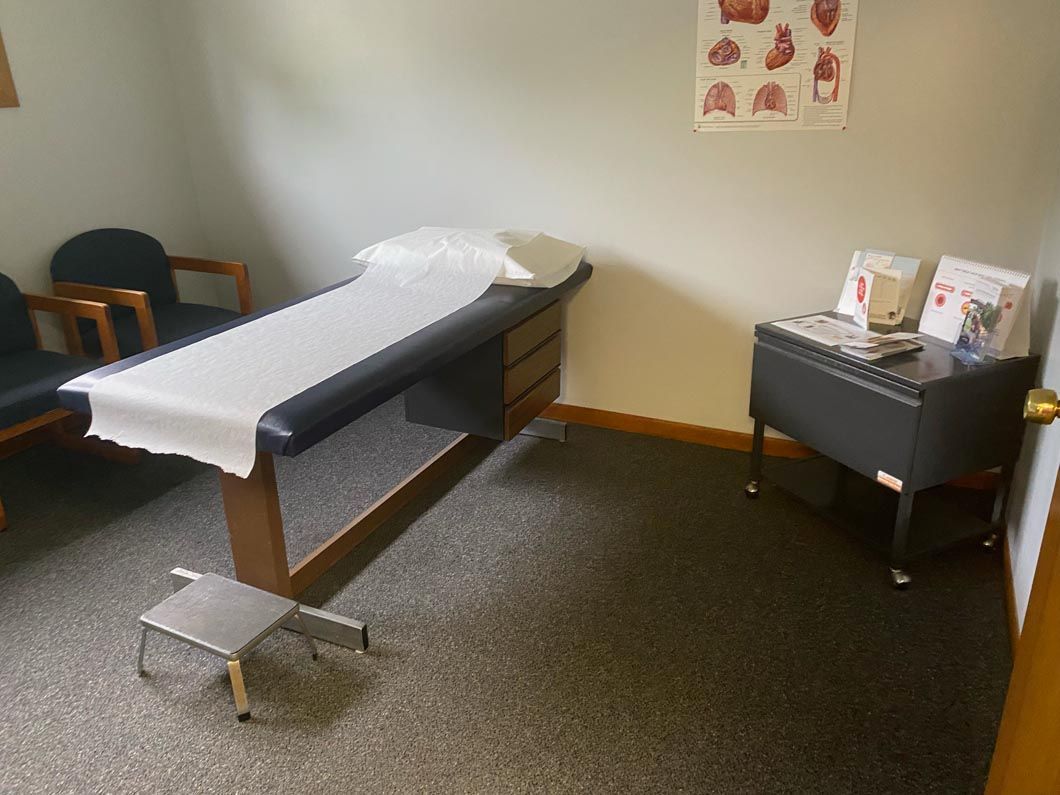 An exam room with a black exam table, paper cover, step stool, chairs, and a medical poster on the wall.