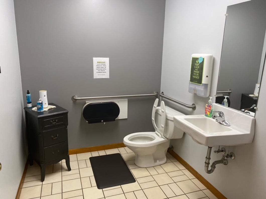 A modern, accessible restroom with a white toilet, grab bars, sink, mirror, and small cabinet against grey walls.