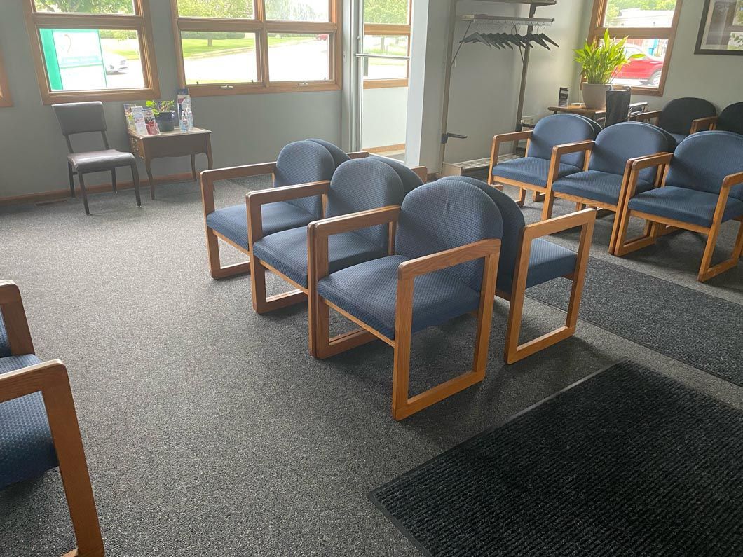 A waiting room with rows of blue chairs with wooden frames, arranged on gray carpet in front of large windows.
