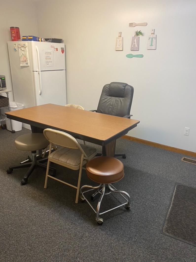 A breakroom with a rectangular table, an office chair, a folding chair, two rolling stools, and a white refrigerator.