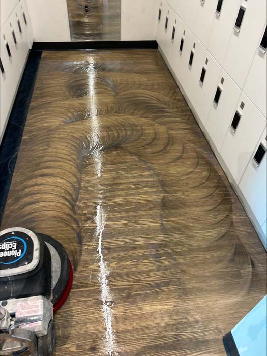 A floor buffer machine on a wood-look floor in a locker room, creating a series of circular patterns during cleaning.