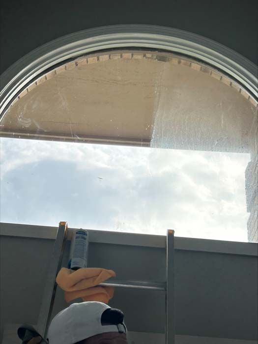 A person on a ladder cleaning a large, arched window against a cloudy sky.