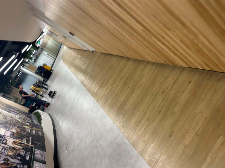 A person in athletic wear stands in a modern hallway with light wood flooring and walls, illuminated by bright overhead lights.