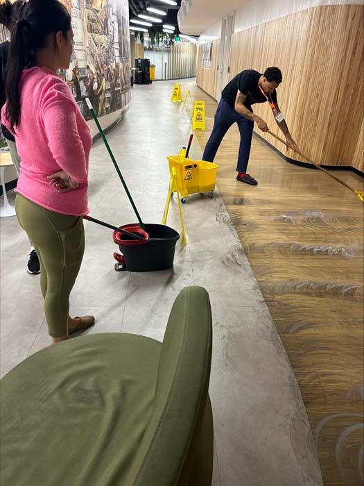 A person mops a wood-paneled hallway while another individual stands nearby with a cleaning bucket and equipment.