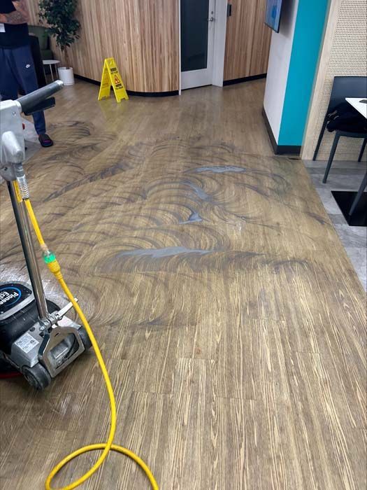 A floor scrubber cleans a patterned wood-look floor in a commercial space, leaving circular marks on the surface.