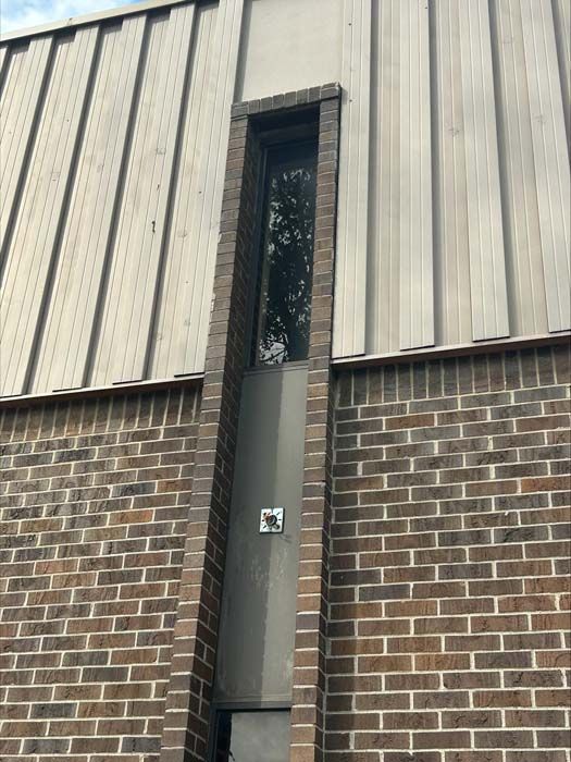 A vertical, narrow window set into a brown brick facade, bordered by vertical metal siding above and on the sides.