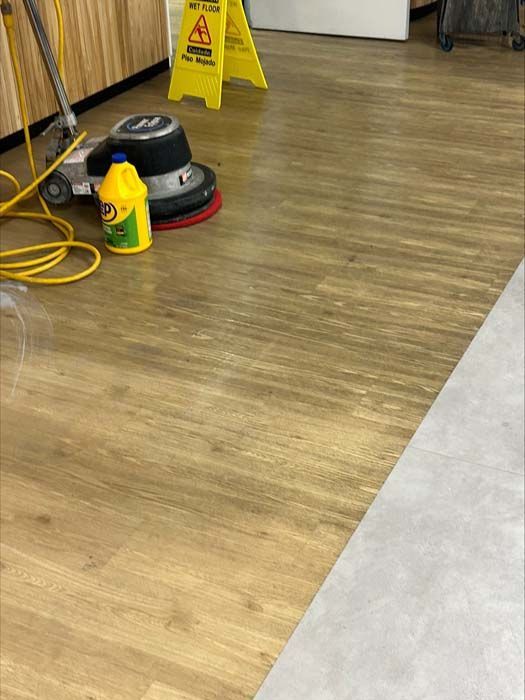 A commercial floor buffer and a yellow bottle of cleaning solution sit on a wooden-look floor near a wet floor sign.