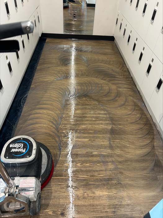 A commercial floor buffer machine cleans a wood-grain hallway floor, leaving circular scrub patterns on the wet surface.