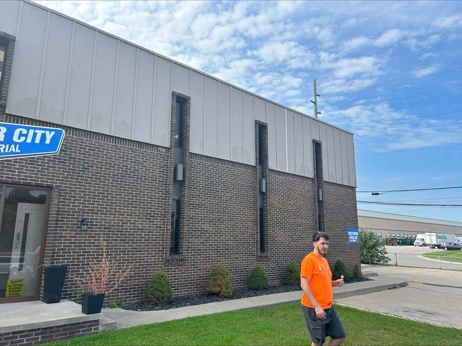 A person in an orange shirt stands on a lawn next to a brick building with white metal siding under a blue sky.