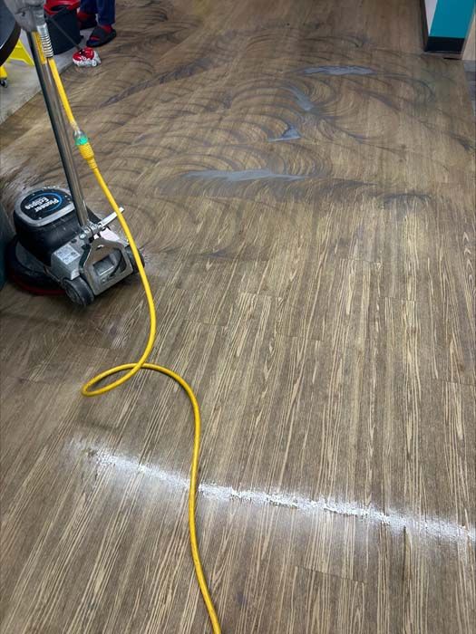 A floor buffer machine cleans a wood-look vinyl floor, showing a path of cleaned surface and swirling dirty residue.