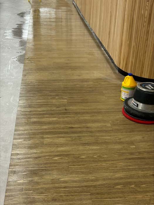 A wood-look floor being buffed, showing a shiny cleaned section beside a scrubbed area with a floor buffer and soap.