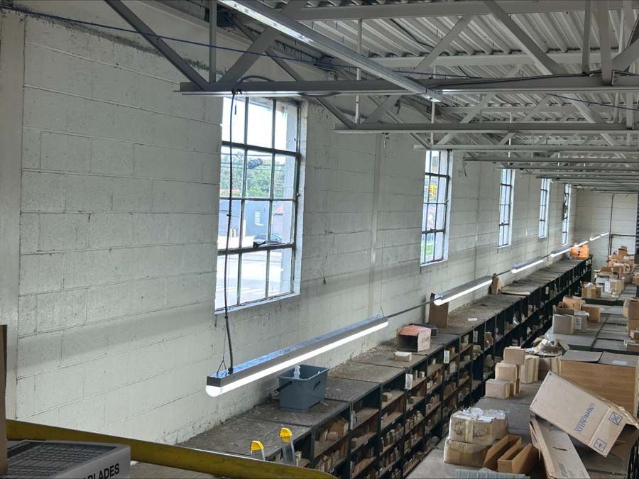 Long, industrial storage room with white brick walls, grid windows, overhead lights, and metal shelving filled with boxes.