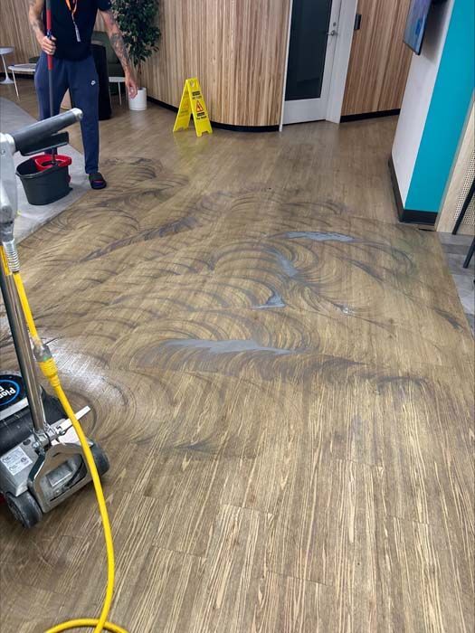 A worker uses an industrial floor scrubber in a room with wood-look flooring and a yellow 